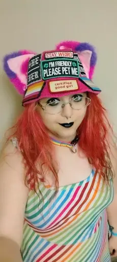 selfie of me vanta enby girl extraordinaire. i have hot pink hair and i'm wearing black eyeliner and black lipstick and my hot pink cat ear bucket hat with patches sewn on and a rainbow collar and rainbow dress and smiling happily