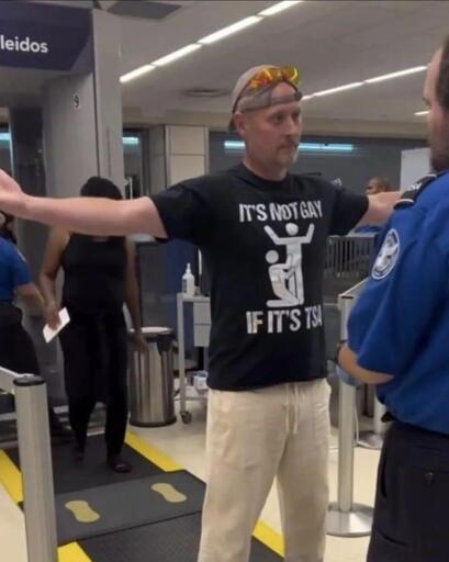 Man preparing for getting searched by the TSA, while wearing a shirt with the words “It’s not gay if it’s TSA” and a picture of 1 man on his knees, touching another one standing up.