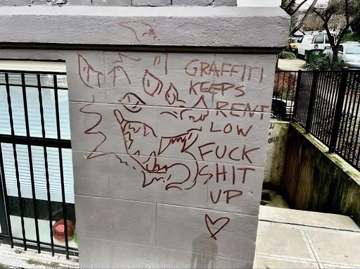 The streetside corner of an apartment building is pictured with linework graffiti of a smiling, three eyed, horned dog demon or dragon with monstrous teeth and its tongue lolling out. Next to the animal is the capitalised writing “graffiti keeps rent low fuck shit up” and a heart symbol.