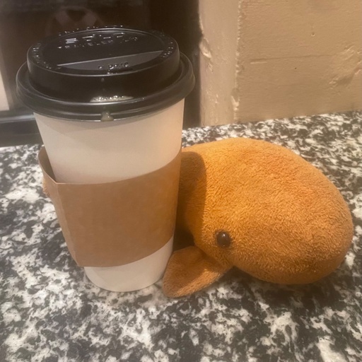 Orange-colored sperm whale plushie doll, next to a paper “to go” cup of coffee, on a marble table top.