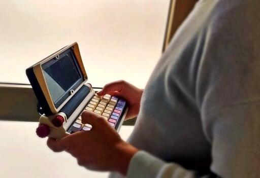 A person is holding a small retro laptop-like computer. It's looks like a stylish old-school word processor.