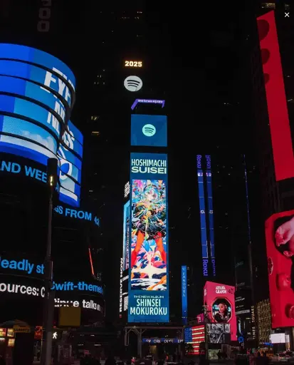 A photo of Times Square taken from the street. An ad for Hoshimachi Suisei's new album Shinsei Mokuroku is prominently displayed on one of the digital billboards.