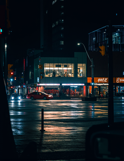 A nighttime scene of an urban street corner featuring a modern two-story building with large glass windows, illuminated from within, showcasing a brightly lit interior. To the left of the building, there is a red car making a right turn on the wet street. The background includes tall, dark buildings, illuminated by a cool, bluish artificial lighting.